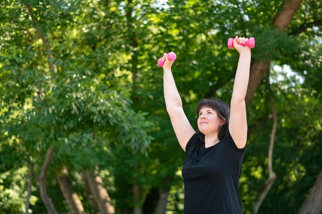 Young girl performs exercises for the hands with the help of dumbbells. Outdoor exercise. Fitness in the park, healthy lifestyle