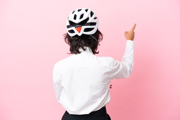 사진 집게 손가락으로 뒤를 가리키는 분홍색 배경에 격리된 자전거 헬멧을 쓴 어린 소녀