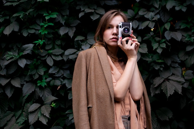 Молодая девушка-фотограф стоит с пленочной камерой возле стены из листьев в лесу, женщина фотографирует на природе