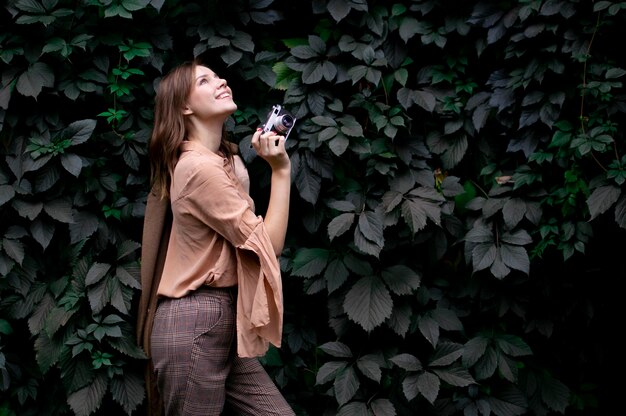 Young girl photographer stands with a film camera near a wall of leaves in the forest