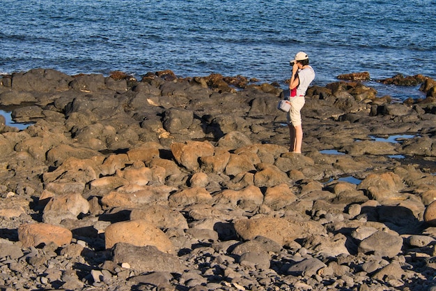 Young girl photographing the rugged terrain beach formed by volcanic rocks from cooling lava