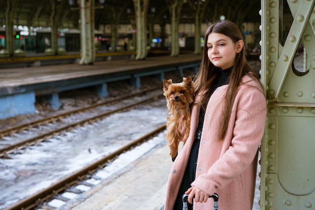 Young girl in pink coat at train station beautiful woman stands on platform with suitcase and a dog ...