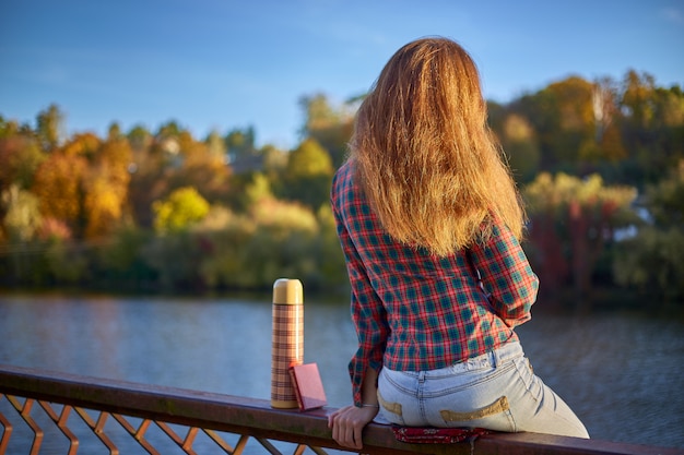 Young girl in plaid shirt sitting on iron railing at river embankment