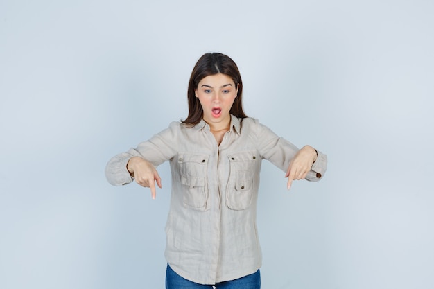 Young girl pointing down with index fingers in beige shirt, jeans and looking surprised. front view.