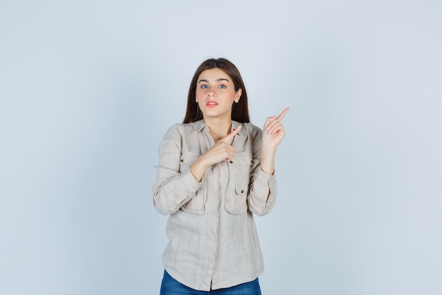 Young girl pointing upper right side with index fingers in beige shirt, jeans and looking cute. front view.