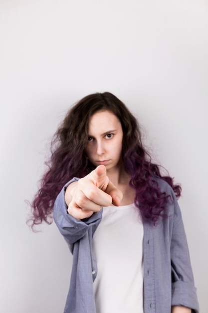 Young girl points finger in lens, serious face. Coloring purple hair. Gray wall