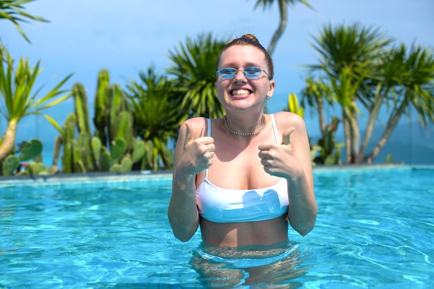 Молодая девушка в бассейне показывает большой палец вверх и улыбается в отпуске летом в теплую погоду