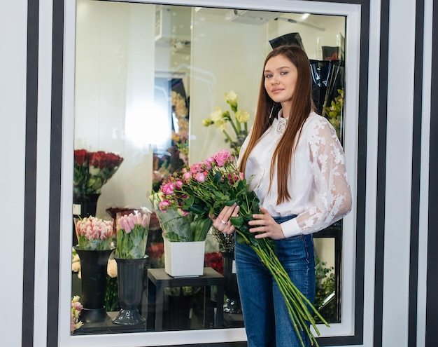 Una giovane ragazza posa con un bellissimo bouquet festivo sullo sfondo di un accogliente negozio di fiori fiorai e bouquet in un negozio di fiori piccole imprese