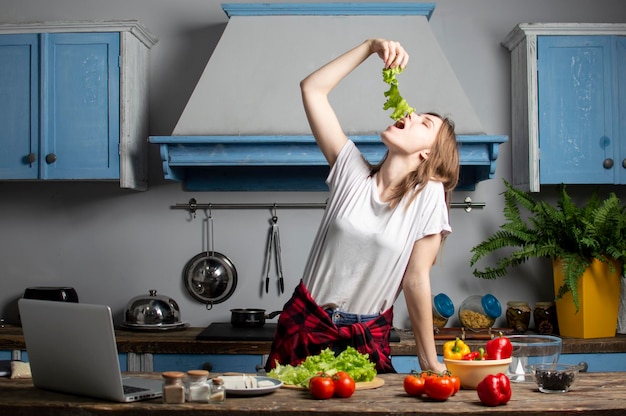 Молодая девушка готовит вегетарианский салат на кухне, смотрит в ноутбук и ест зелень