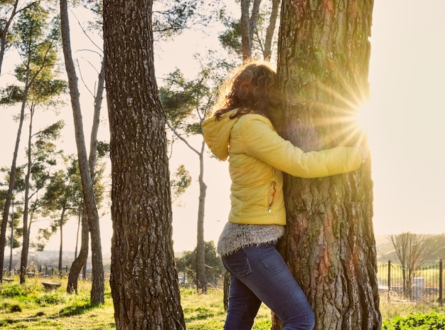 Young girl in profile with yellow jacket and blue pants hugging a tree in a pine forest at sunset with a solar star peeking from the right side of the tree