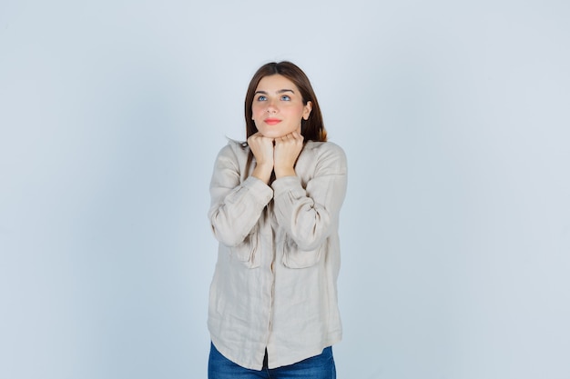 Young girl propping chin on fists, thinking about something in beige shirt, jeans and looking pensive , front view.