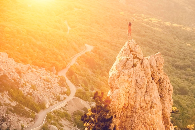 The young girl raised her hands up towards the sun at the peak of a high cliff on the background of mountains