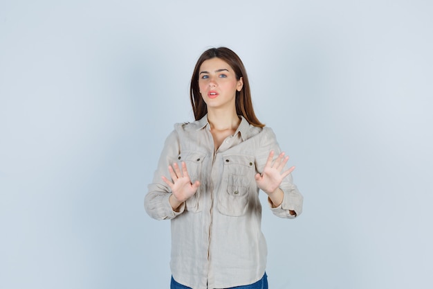 Young girl raising hands to stop in beige shirt, jeans and looking serious. front view.