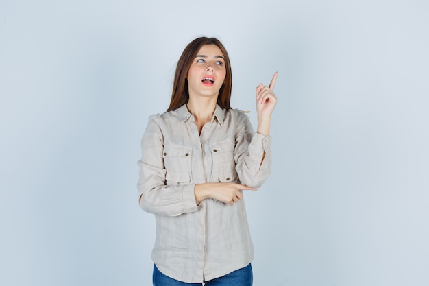 Young girl raising index finger in eureka gesture in beige shirt, jeans and looking sensible , front view.