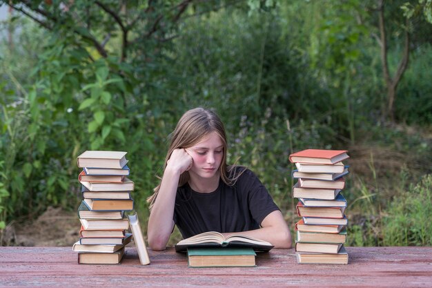 Молодая девушка читает книгу в саду за деревянным столом со стопкой книг. Концепция домашнего образования