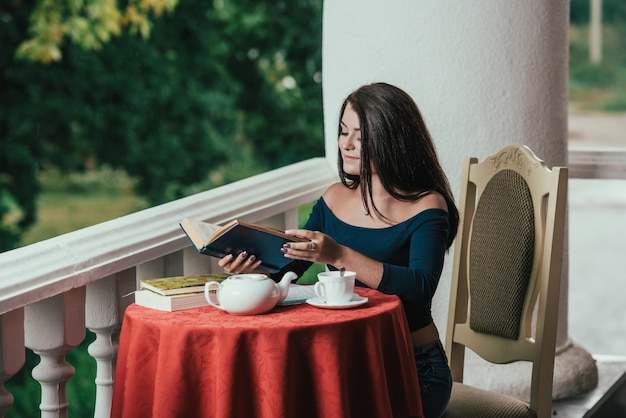 young girl reading book while drinking coffee at sunny day sitting on the balcony .