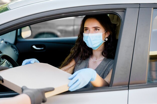 若い女の子は、医療マスクと手袋で車の中でピザボックスを受け取ります