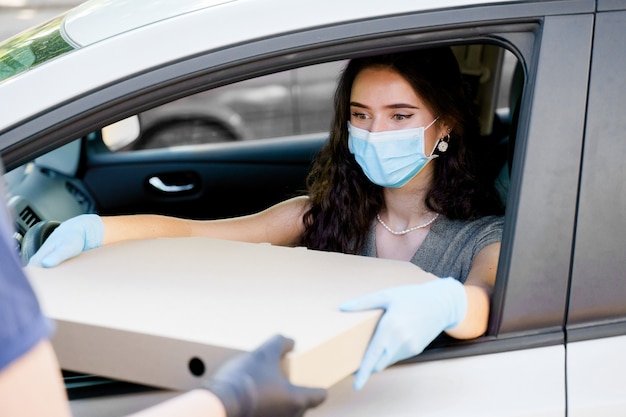 若い女の子は、医療マスクと手袋で車の中でピザボックスを受け取ります