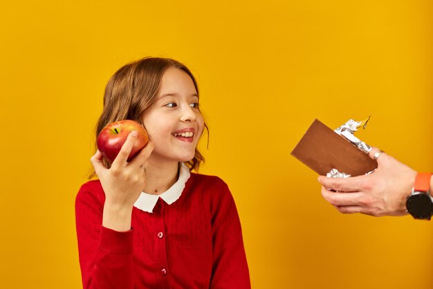 Young girl in red choosing an apple over chocolate against a yellow background