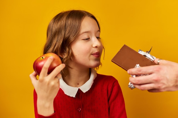 Giovane ragazza in rosso che sceglie una mela sul cioccolato contro uno sfondo giallo