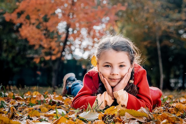 Young girl in a red coat lies on the grass and leaves in an autumn park