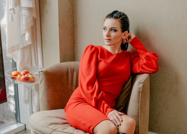 Young girl in a red dress indoors. model. Emotions of a modern lady. The girl is sitting in a chair