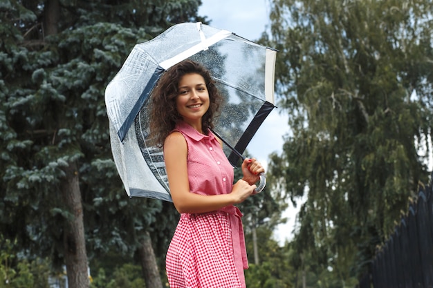 Young girl in a red dress with a transparent umbrella dancing in the rain standing in a puddle