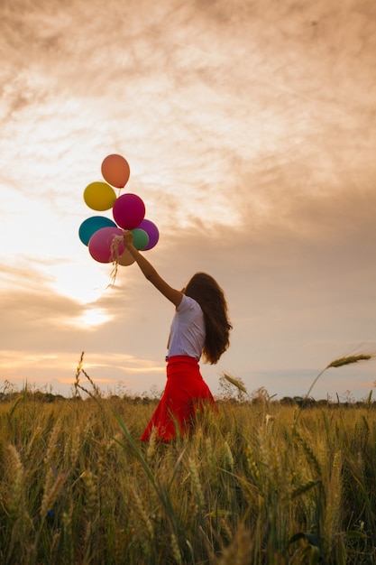 Young girl in a red skirt running with colored balloons in wheat field. Nature inspiration, backlight