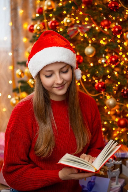 Giovane ragazza in maglione rosso e cappello da babbo natale legge un libro durante le vacanze invernali vicino all'albero di natale colori caldi e luminosi felice atmosfera magica festosa interni