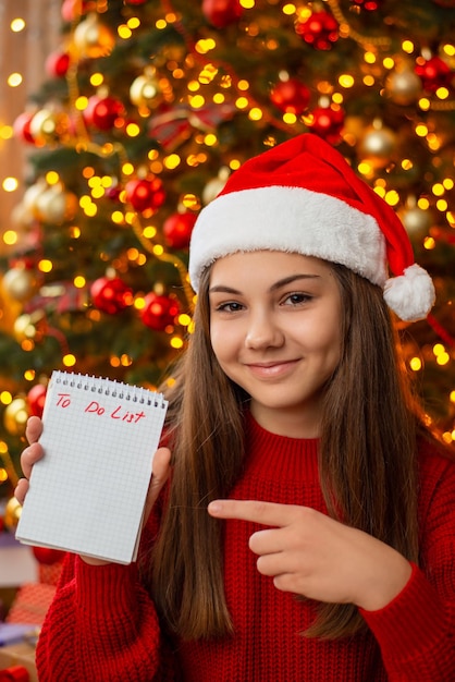 Young girl in red sweater and santa hat with big smile on her face holds notebook
