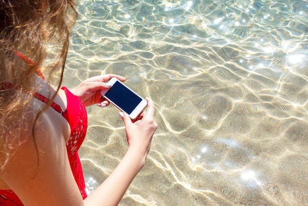 빨간 수영복을 입은 어린 소녀가 휴가 해변 컨셉에서 휴대폰으로 바다 사진을 찍고 있다