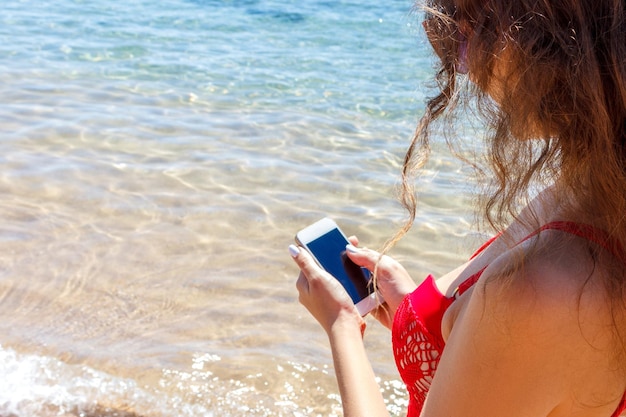 빨간 수영복을 입은 어린 소녀가 휴가 해변 컨셉에서 휴대폰으로 바다 사진을 찍고 있다