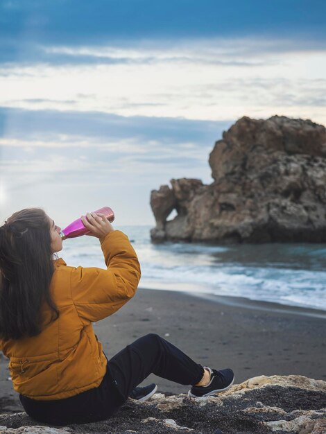 Young girl relaxing on a beach at sunrise looking at a rock drinking from a bottle Cool day with clouds Orange and blue colors Vertical shot