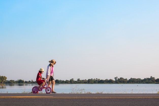 Young girl ride bicycle with mother helping on road with river background 