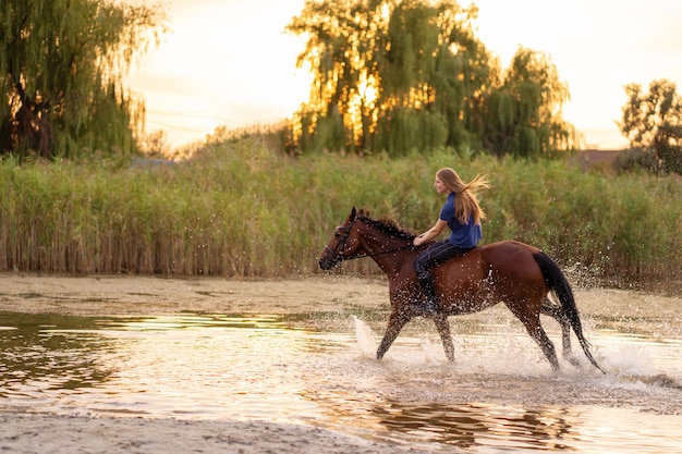 浅い湖で馬に乗る少女。馬は日没時に水の上を走ります。馬と一緒に気をつけて歩いてください。強さと美しさ