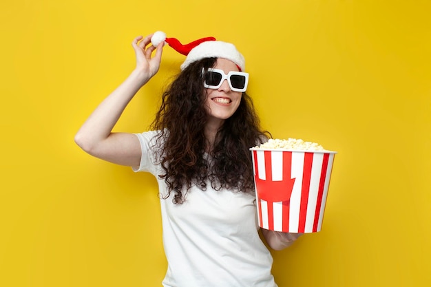 Young girl in santa hat and 3d glasses holds popcorn and smiles on yellow isolated background