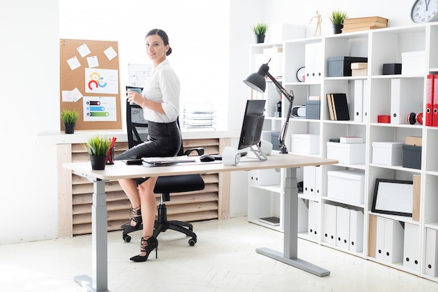 Una ragazza si sedette su un tavolo in ufficio, tenendo una tazza e lavorando con un computer e documenti