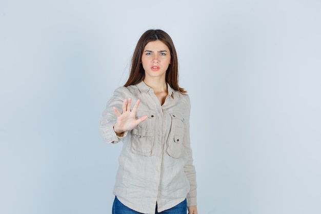 Young girl showing stop sign, grimacing in beige shirt, jeans and looking angry. front view.