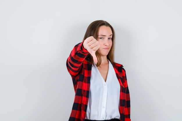 Young girl showing thumb down in checkered shirt, blouse and looking sly , front view.