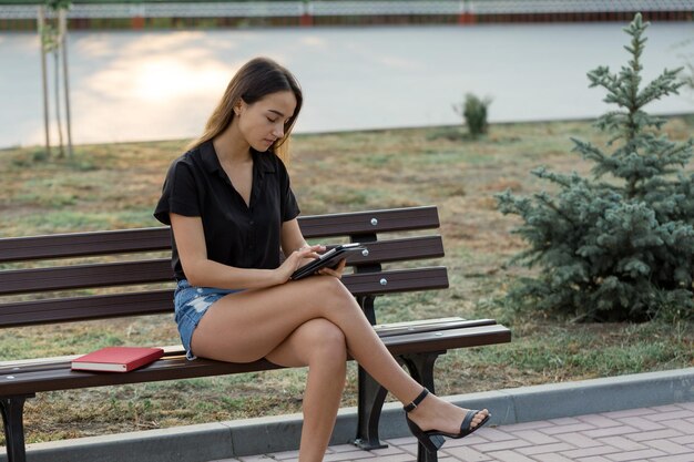 若い女の子が公園のベンチに座ってメモをとるフリースタイルの服を着て顧客と電話で話しているビジネスウーマン