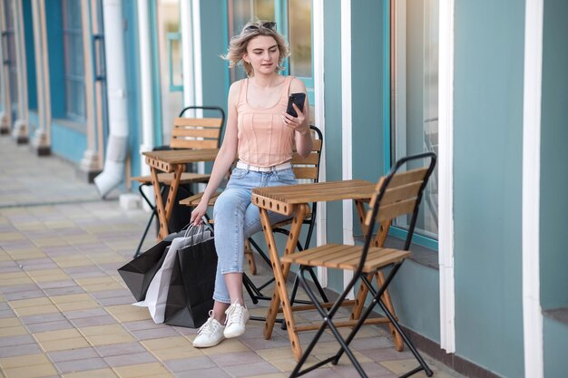 A young girl sits next to a cafe with a phone in her hands With shopping bags