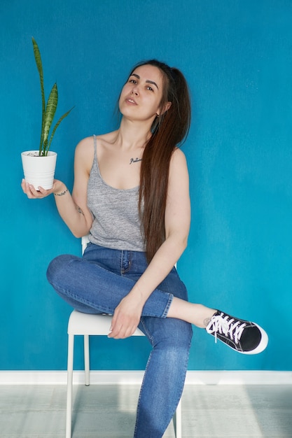 A young girl sits on a chair and holds a flower in a pot in her hands, on a blue background