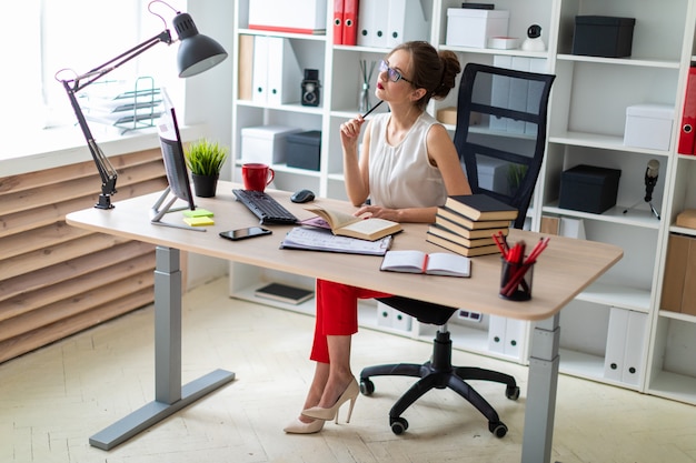 A young girl sits at a computer desk and holds an open book and a pencil in her hands.