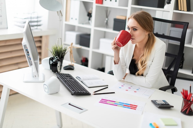 Photo a young girl sits at a computer desk in the office and holds a red cup in her hand.