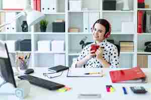 Photo a young girl sits in headphones at a table in the office, holds a red cup in her hands and smiles.