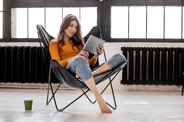 A young girl sits at home in a stylish black leather chair, holds a cup of coffee and a tablet in her hand. Work and relaxation at home