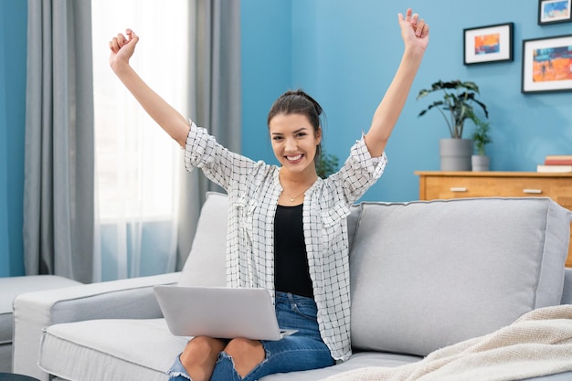 A young girl sits in the living room on the couch with a laptop in lap raises her hands up