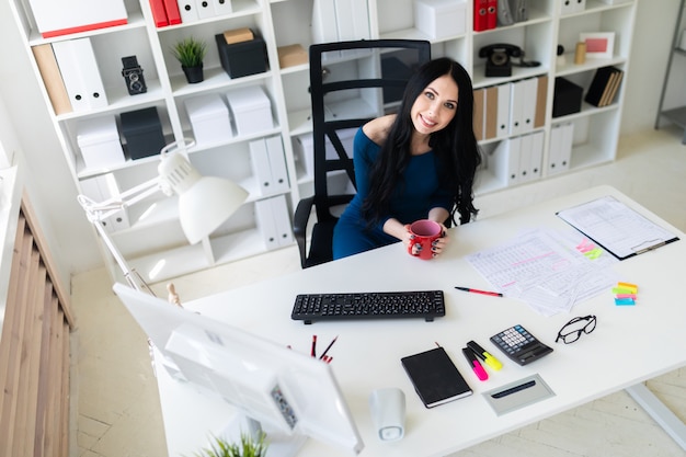 A young girl sits in the office at the table and holds a red cup in her hands