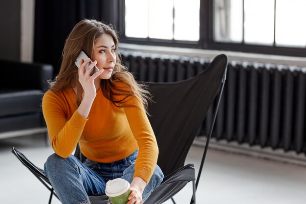 A young girl sits in a stylish black leather chair, holds a cup of coffee and a phone in her hand. Work, communication, relaxation at home