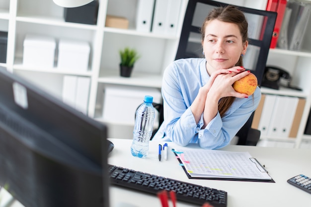 A young girl sits at a table in the office and holds an apple in her hand.